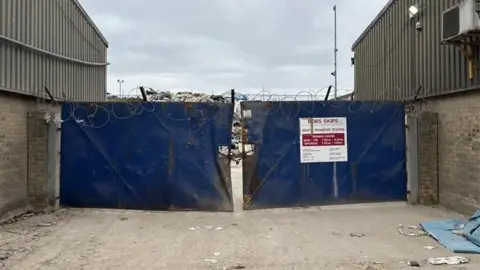 Jodie Halford/BBC A blue gate in front of a waste management site. There is barbed wire at the top and the gate is padlocked shut. There is a white sign on one panel of the gate with red lettering showing the name of the firm Bobs Skips and its opening hours underneath.