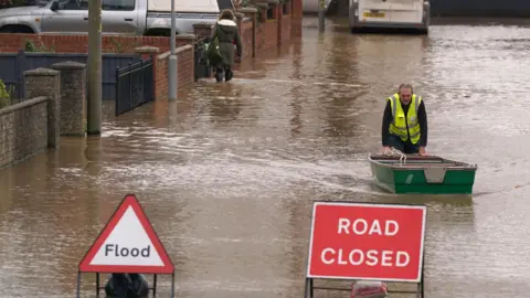 A man in a hi-vis tabard pushes a green rowing boat through floodwater in the middle of a residential street. Behind, a woman is walking in calf-high water at the side of the road. In the foreground, signs reading Flood and 'road closed' are at the edge of the water.