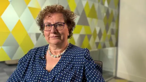 Dame Hilary Cass is sat looking at the camera in a meeting room. She is in front of a glass wall with a geometric pattern.