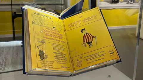 Leeds Central Library A book in a glass cabinet open on a spread of yellow pages. On the right hand page is a drawing of a man with a bowler hat, umbrella and brief case in a Bradford City shirt.