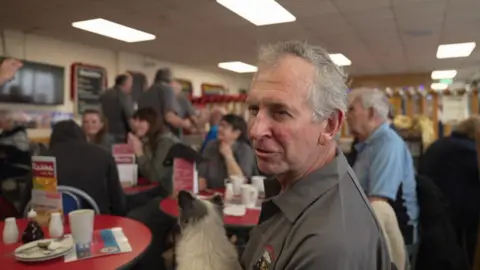 Andrew sits in a room with lots of other people in the background and has a little dog on his lap.