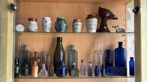 Eddie Goodall A wooden display cabinet holds two glass shelves filled with vintage bottles and ceramic jars. The top shelf includes old dairy and medicinal containers with printed labels, while the bottom shelf shows glass bottles in different shapes and colours.