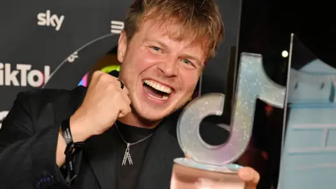 Getty Images Max Klymenko raises a fist in celebration while smiling happily and holding an award which is the shape of the TikTok logo against a background of advertisements at the awards ceremony in London on Thursday.