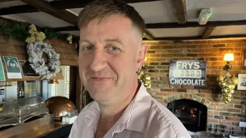 BBC / Phil Harrison A man with grey hair and a pink shirt stands in front of a wooden pub bar and a brick fireplace. 