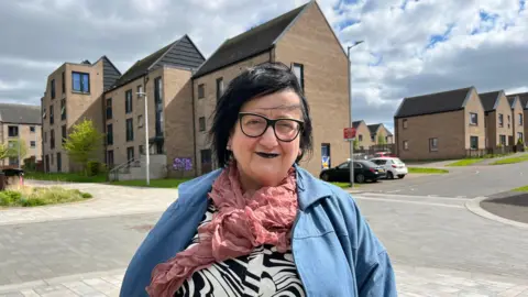 Elaine Ellis - a woman with dark hair, wearing a white an black top with blue jacket and pink necktie. 