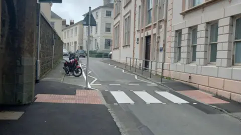 States of Guernsey A zebra crossing on a single-lane road in Guernsey surrounded by buildings, with two cars in the distance and a parked motorbike on the left