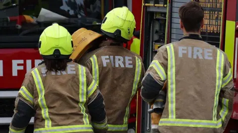 Generic image of the backs of three firefighters walking towards an engine, the two to the left are wearing helmets 