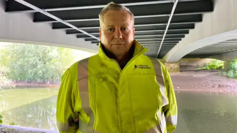 Terry Robinson. He is pictured underneath the A40 Over Bridge in Gloucestershire. He has short grey hair and is wearing a yellow high vis jacket with a National Highways logo on the chest. He is looking at the camera and standing with his hands behind his back.