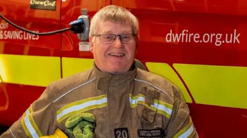 Keith Prince smiling for a picture with his firefighter uniform in front of a Dorset and Wiltshire rescue fire engine.