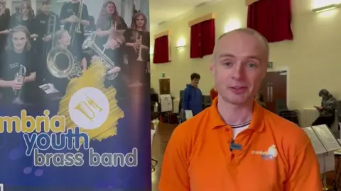 Stuart Humphries is wearing an orange t-shirt stands in a rehearsal room in front of a blue and yellow banner - "Cumbria Youth Brass Band" - featuring images of young people playing brass instruments. 