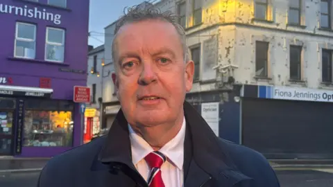 Michael is looking at the camera. He has grey hair. He is wearing a white shirt an red and navy tie. He is also wearing a navy coat.