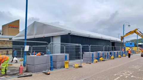 Shot of the temporary market being installed by workers. The building is a large grey building, with a V-shaped roof. Construction workers and fencing surrounds it.