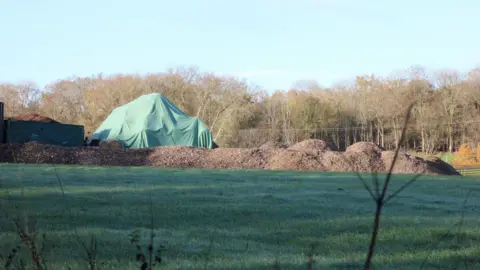 A large green tarpaulin covers a mound of materials on a field, with several piles of brown waste nearby. Trees line the background.