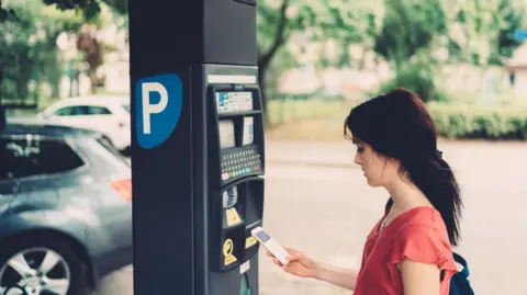 A young woman in a red top, using her phone next to a parking meter