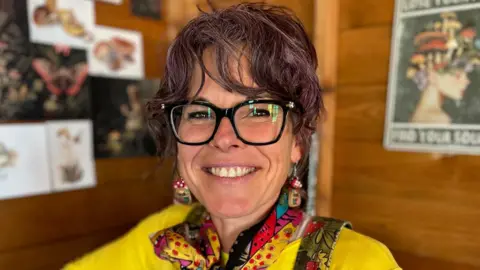 Kylie Dixon is pictured seated inside her allotment shed. She is wearing dark rimmed glasses, a yellow t-shirt and green dungarees with a brightly coloured neck-scarf and mushroom-shaped earrings. She has purple-tinted dark curly hair that is tied up. 