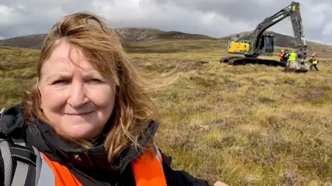 Foto estilo selfie de PA de Lorna Dawson, que tiene el pelo largo y castaño. Viste una chaqueta negra y un chaleco reflectante naranja. Al fondo se ven páramos y colinas y al fondo se ve una gran excavadora.