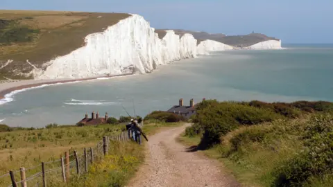 Jeff Overs/BBC The huge white cliffs of the Seven Sisters and the shoreline beneath them with a path leading to the coastguard cottages with two anglers walking down towards the beach