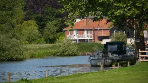 Shaun Whitmore/BBC A boat anchored on a river close to the riverbank. There is a house in the background on the other side of the river. There are trees and long grass areas flanking both banks. 