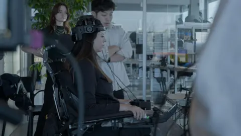 Dentsu In a lab, Breanna looks at a laptop on a desk while holding the arms of her wheelchair and concentrating on using the brainwave interface. Two researchers stand by her side, looking at the screen. 