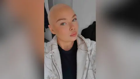 Ellie Philpot A girl, Ellie, with makeup on, a black t-shirt and white marble-effect hoodie. She looks glamorous as she poses for a selfie with a shaved head. 