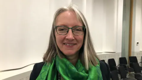 Vikki Irwin/BBC Caroline Topping has blonde hair, is wearing black glasses and a green scarf and is smiling at the camera.