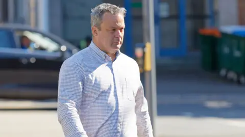 A man with grey hair walking. He has a white checked shirt on. 