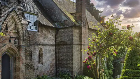 Ewelme Almshouse Charity A beautiful brick building with gothic arches and a wooden door off to the left. Through a rose arch on the right you can see a topiary ewe bush. The photo is bathed in a heavenly glow of sunlight.