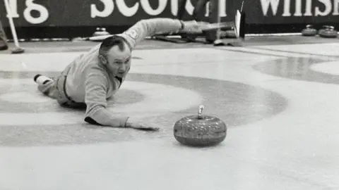 McMillan family Hammy McMillan lying on his stomach on the ice with his right arm resting on the ice, having just thrown the curling stone- his left arm is held in the air