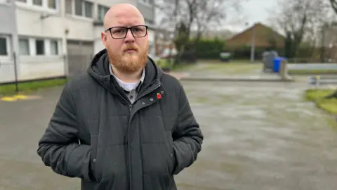 Josh Kearns standing outside a tower block. He is wearing a body warmer, a black hoodie and black trousers. He has a bald head and his hands are in his pockets. 