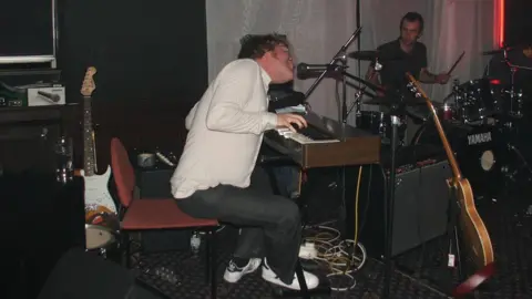 Andrew Woodger/BBC Mr Simpkins wearing a white shirt, black trousers and white shoes behind a piano. He is singing into a microphone with a drummer in front of him and a stray guitar.