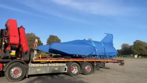 The Bluebird concealed under a blue covering, strapped on top of the trailer of a truck.