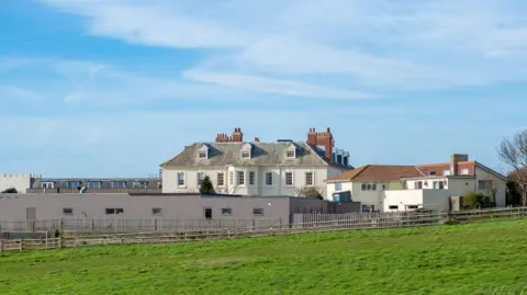 Getty Images/Wirestock A low-angle of Moonfleet Manor hotel seen from SW Foot path grass around cloudy sunlit sky background