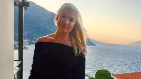 Family photo Bronwen is pictured on a balcony in a black off the shoulder dress on holiday with the sea and sunset behind her. She has long blonde hair and is smiling at the camera