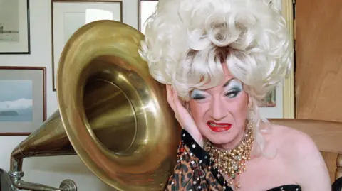 Paul O'Grady as his alter ego Lily Savage in a blonde wig next to a grammarphone 