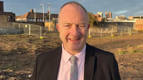 Head and shoulders picture of Cllr Ray Morgon, leader of Havering Council, on an empty plot of land in Romford that is waiting to be developed