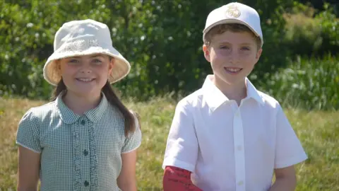 Jamie Niblock/BBC A young girl and boy smile at the camera. They are both wearing school uniform and sun hats and are standing against a backdrop of grass and a riverbed.