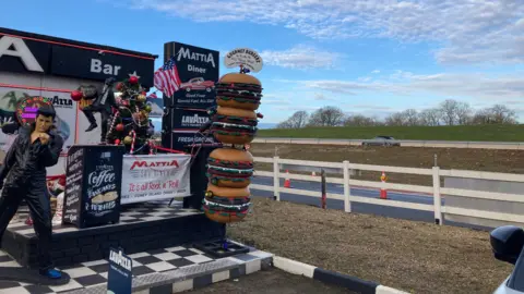 On the left is the outside facade of the diner, with a life-size Elvis Presley statue, and a stack of overly large burgers. A fence runs between the building on the road on the right. On the road in the background a car is driving past.