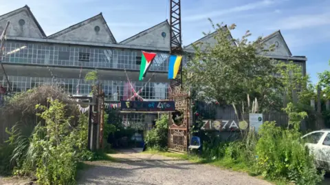 A former factory building with an angular roof and overgrown greenery. The flags of Ukraine and Palestine fly at the front of the building