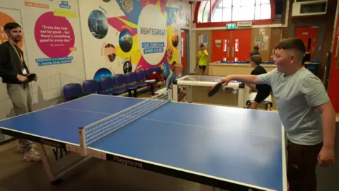 A man wearing a lanyard and a young boy, Harvey, playing table tennis on a blue table tennis table. Harvey is hitting the ball with his bat reaching towards the man.