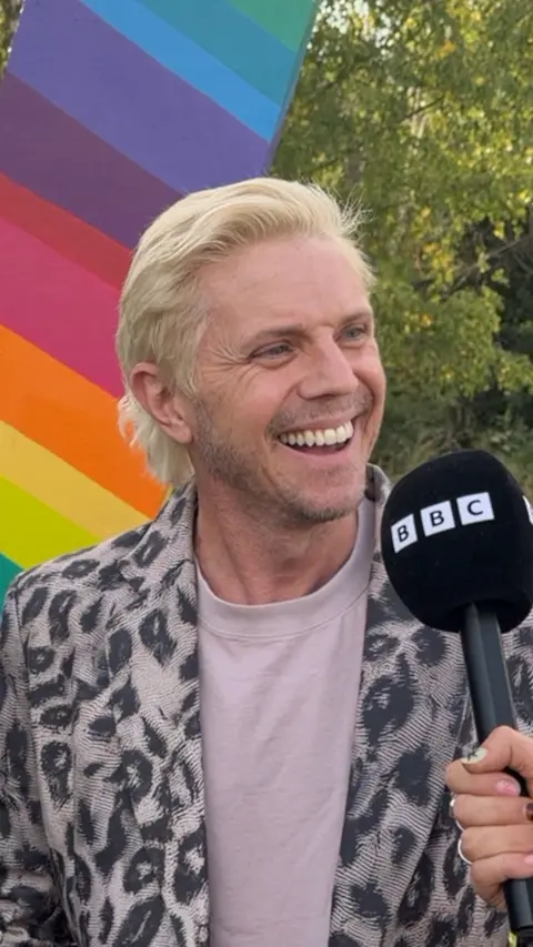 Jake Shears of Scissor Sisters smiles during an outdoor interview with a BBC microphone. He is wearing a black-and-white patterned jacket over a light-coloured top. A large rainbow-coloured backdrop and green trees are visible behind him.