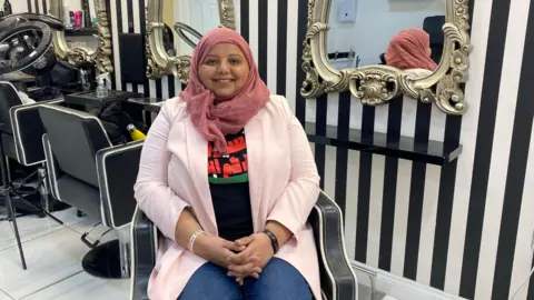 Shaista is sat in a salon with black and white striped walls and ornate mirrors. She wears a pink hijab, a lighter pink jacket with a black shirt underneath, and jeans.