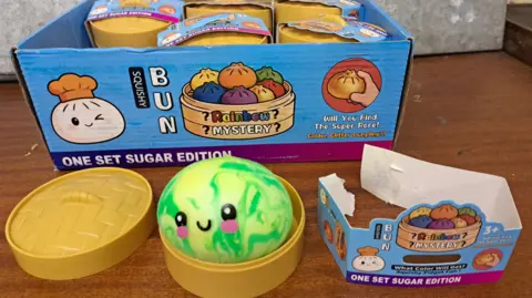A green squishy ball with a light green and darker green swirly pattern, circular black eyes, a black curved line for a mouth and pink circles for cheeks is in a mini plastic container made to look like a bamboo steamer. The packaging for the illegal toy, placed next to it, and also on a larger box of stock behind it, has writing including 'Rainbow mystery', 'Squishy bun', 'One set sugar edition' and 'will you find the super rare golden glitter dumpling?!' on it, with colourful cartoon images of toy squishy buns.