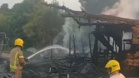 A screenshot from a video showing firefighters putting out the blaze. A charred, mangled building reveals where the wooden pigeon loft used to be. A sketal frame is all that remains, with the rest of the building collapsed into a pile of black debris on the ground. The firefighters, in protective gear and yellow helmets, spray a jet of water towards the site, white steam rising from it.