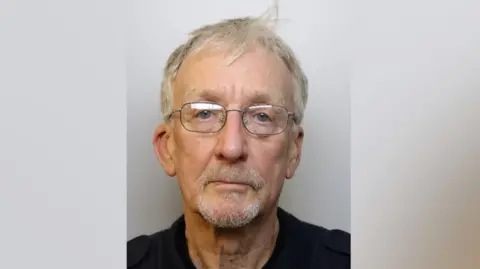 David Fretwell is wearing glasses with thin metal frames. He has wispy pepper and salt hair and has a white beard and a round face. He is wearing a black top.