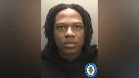A man with black hair, wearing a black hoodie, stands in front of a plain wall in a police mugshot.