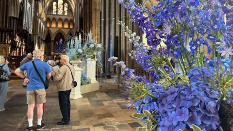 An arrangement of blue flowers in the foreground and in the background on white pillar stands. A few people are standing to the left in the image.