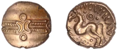 Noonans A gold coin with artwork. One one side there is a drawing that appears to be a horse and other markings including lines and dots are visible. The reverse side show pattern across the middle of the coin.