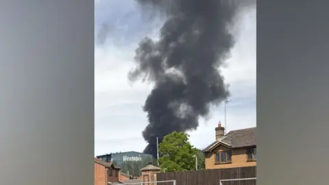 A large plume of thick black smoke rises into the air. There are homes around it and an industrial unit with 'Weetabix' on the side.