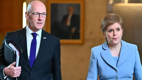 A bald man with glasses and a woman with short brown fair hair stand side by side. The man is wearing a dark suit, white shirt and blue tie. The woman is wearing a light blue suit. There is a painting on a wall behind them, out of focus in the back of the shot. 