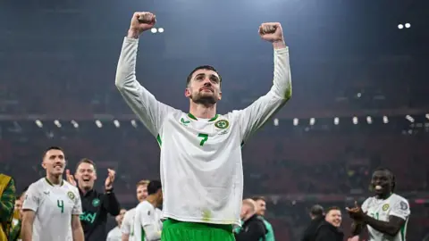 Getty Images Troy Parrott of Republic of Ireland celebrates after the FIFA World Cup 2026 Group F Qualifier match between Hungary and Republic of Ireland at Puskás Aréna in Budapest, Hungary. He has short dark hair wearing his white and green Ireland kit. He is holding up both fists in the air. Other footballers are in the background smiling at him and clapping.
